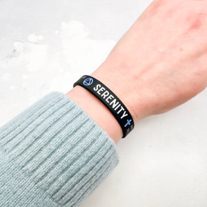 A close-up of a person's wrist wearing a black silicone bracelet with the word 'SERENITY' in white and blue symbols