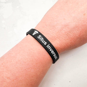A black silicone wristband with white text reading 'Situs inversus' and a small medical caduceus symbol, shown worn on a person's wrist