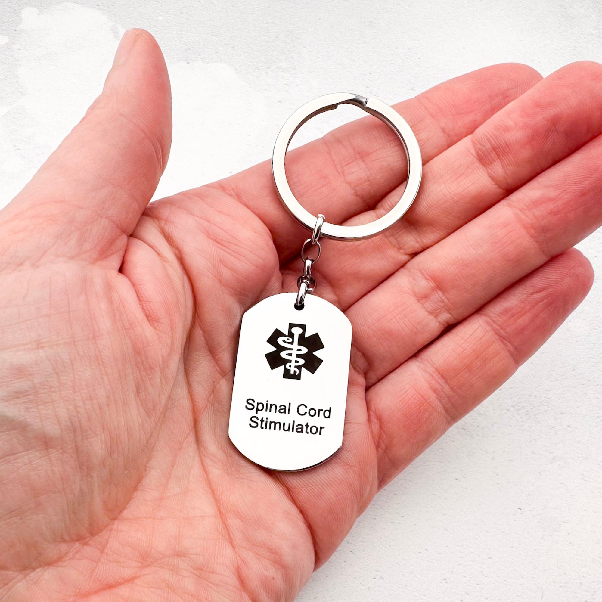 stainless steel keychain with star of life medical symbol and 'Spinal Cord Stimulator' text held in hand