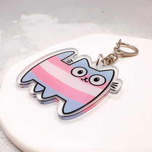A cartoon cat-shaped acrylic keychain with pink, white, and light blue horizontal stripes, transgender