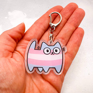 A hand holding a clear acrylic keychain featuring a cartoon cat with transgender horizontal stripes