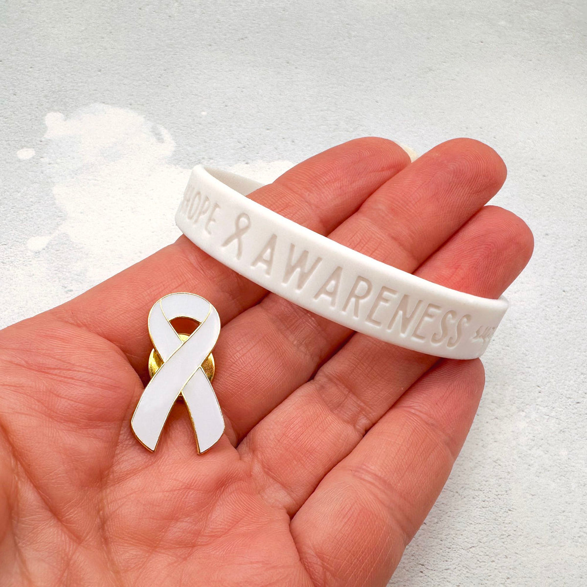 white silicone wristband engraved with ‘Hope & Awareness’ held in hand with matching white enamel ribbon pin