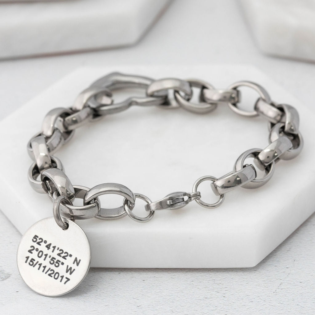 Chunky-style chain bracelet made from stainless steel with heart connector link and a round tag depicting coordinates on a white surface