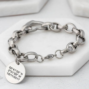 Chunky-style chain bracelet made from stainless steel with heart connector link and a round tag depicting coordinates on a white surface