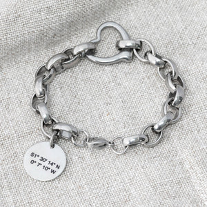 Chunky-style chain bracelet made from stainless steel with heart connector link and a round tag depicting coordinates on a grey background