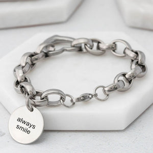 chunky-style stainless steel chain bracelet with a heart connector link and a charm tag with text that reads "always smile" on a white surface. 
