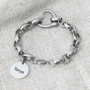 chunky-style stainless steel chain bracelet with a heart connector link and a charm tag with name on a grey textured background. 